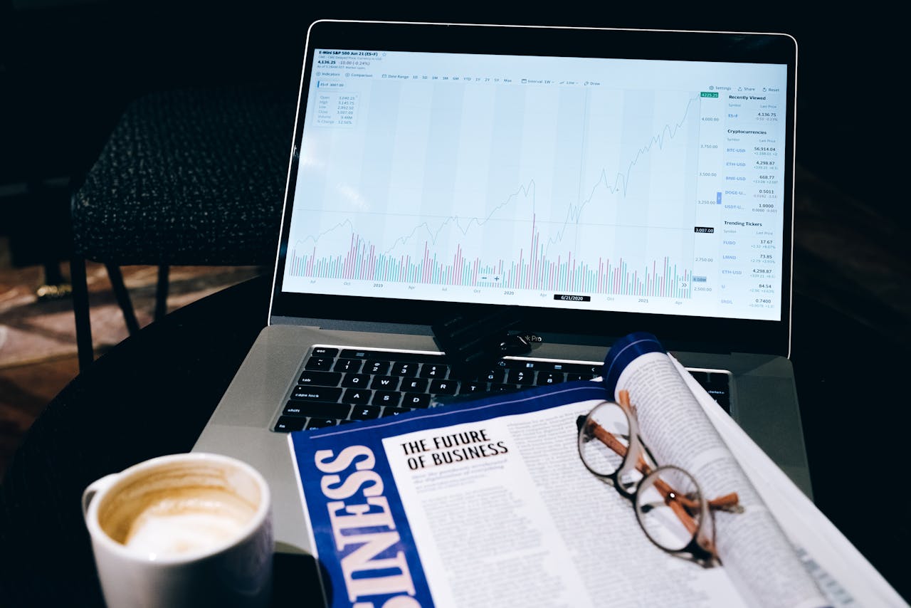 A modern business workspace featuring a laptop showing stock data next to a business magazine and coffee.